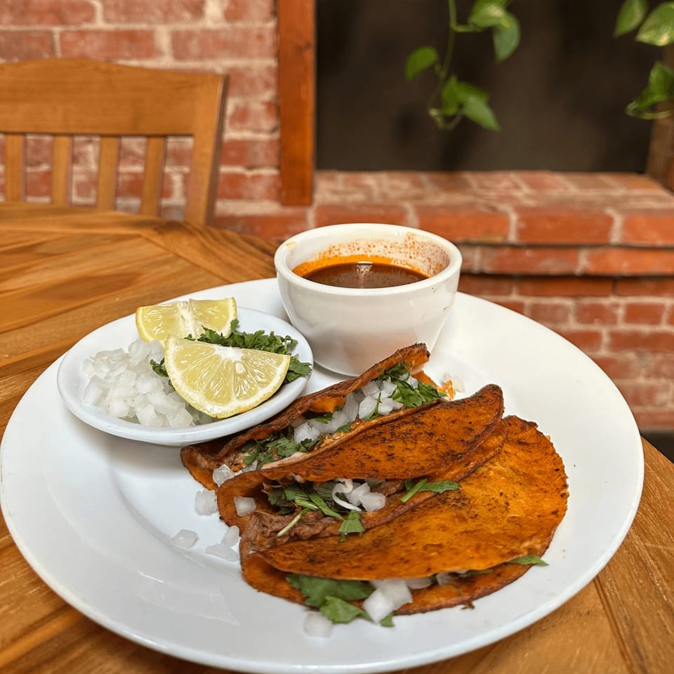 Three birria tacos with cilantro, onions, lime, and a side of consommé on a white plate.