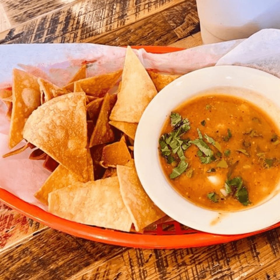A red basket of crispy tortilla chips next to a white bowl of Cocina 1912's salsa on a wooden table.