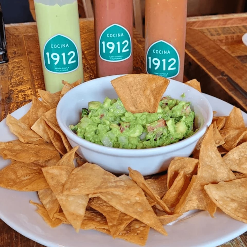 Cocina 1912 guacamole and tortilla chips with three branded sauce bottles on a wooden table.